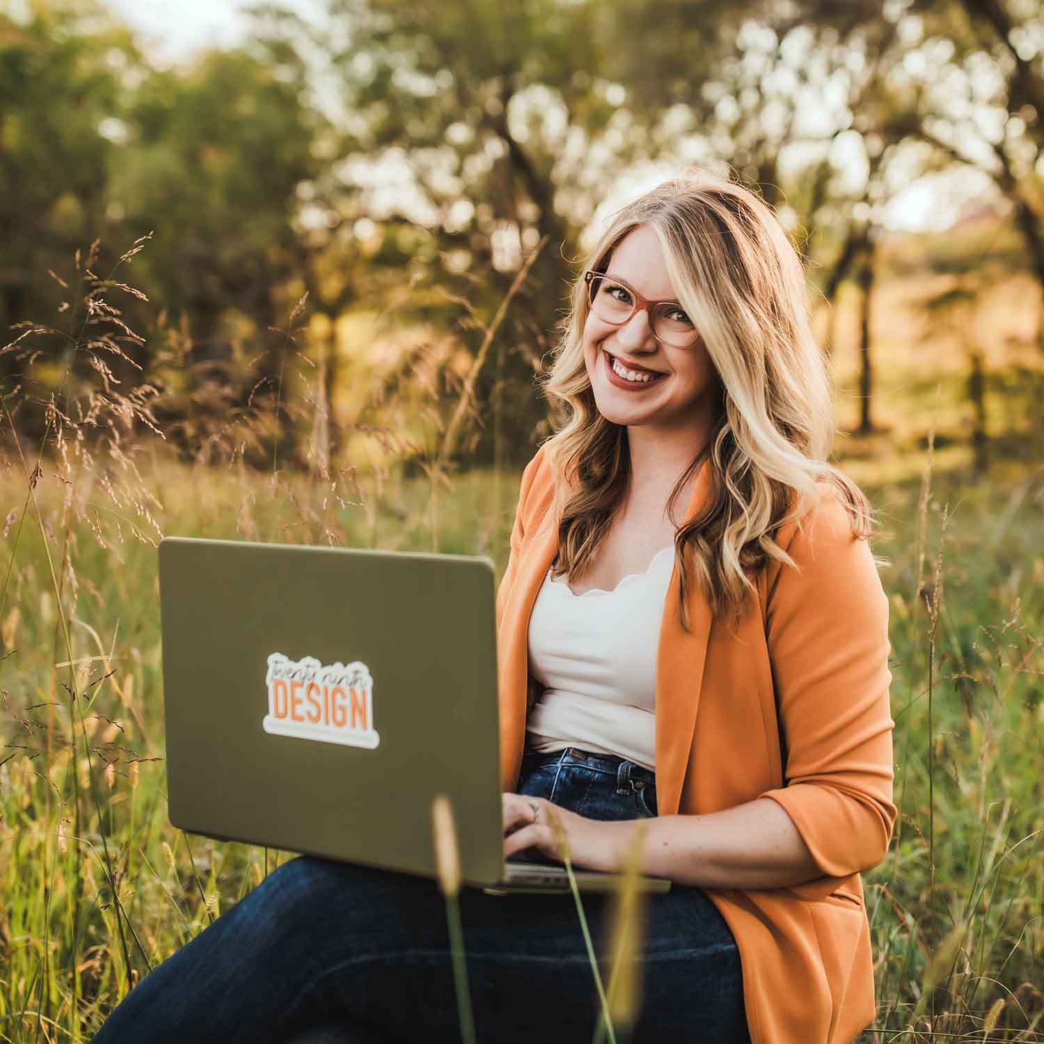 Photo of Hayley Royle, owner of 29th Design, wearing an orange blazer while holder her green laptop open.