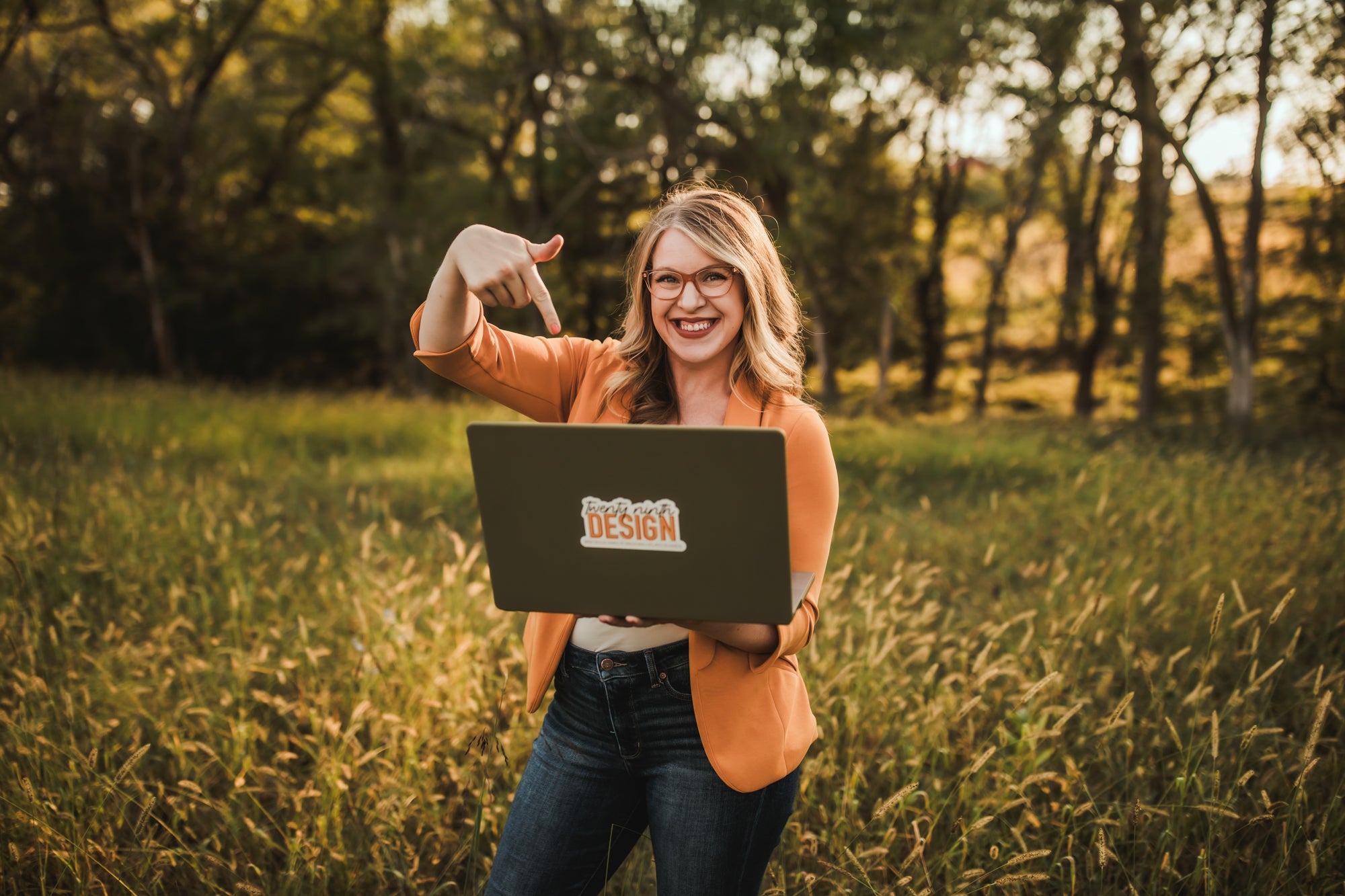 Hayley, owner of 29th Design, standing in a field of grass in the summer while holding her computer with one hand and pointing to it with the other.