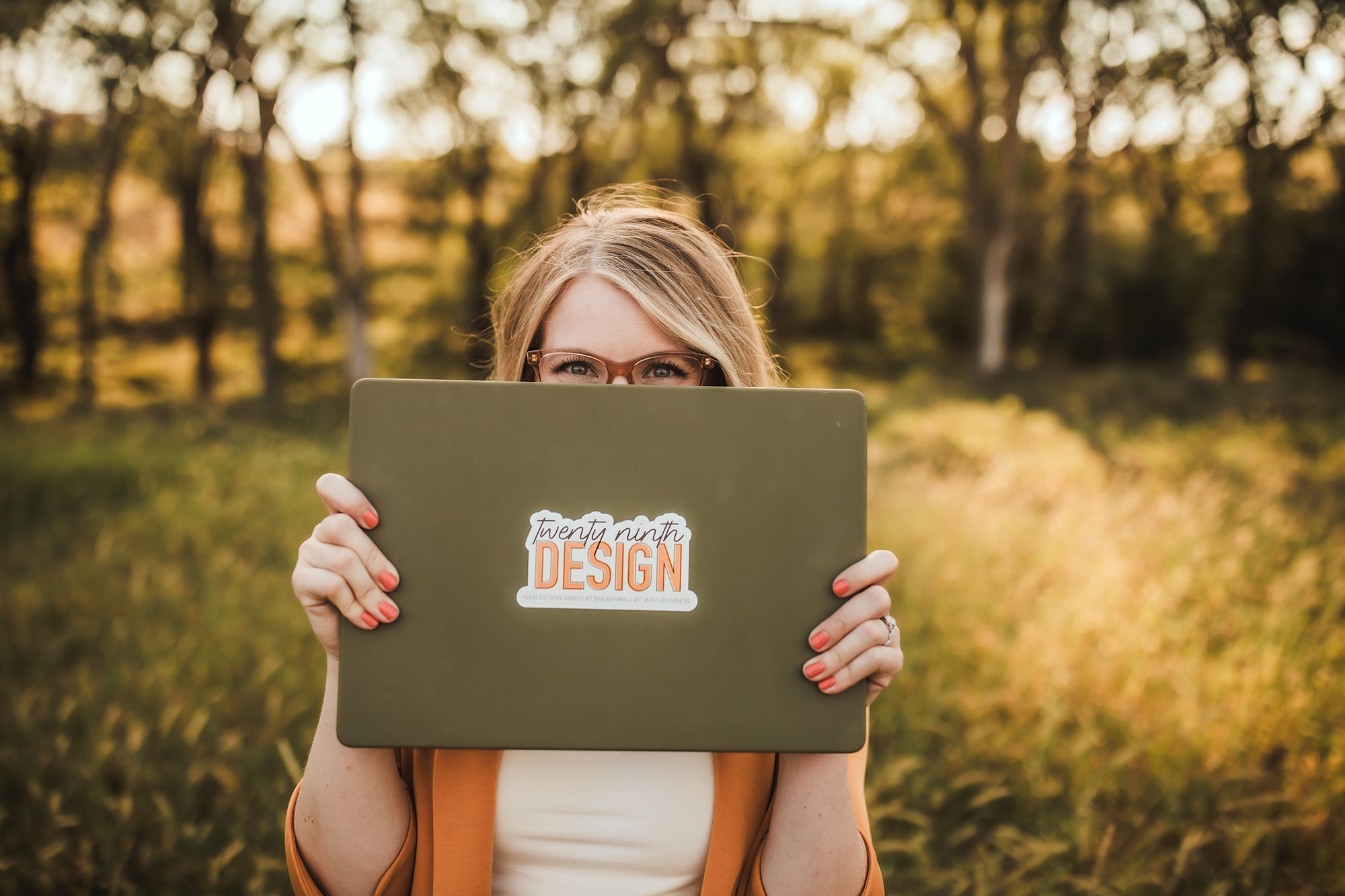 Photo of Hayley Royle, owner of 29th Design, standing in a green Nebraska pasture during golden hour with her green laptop covering her face.  Her eyes are peering over the top of the laptop.